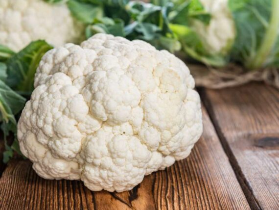 large head of cauliflower sitting on a wooden surface with more cauliflower heads and greens in the background